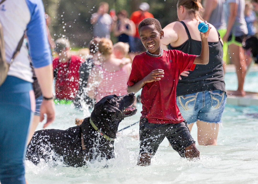 Boy playing ball with his black lab in the middle of a crowded pool both with huge smiles on their faces